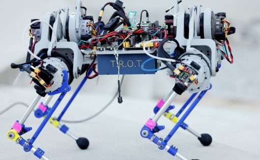 Four‑legged robot with exposed wiring and motors, walking on concrete surface; appears to be a prototype with a blue chassis labeled 'TROT'.