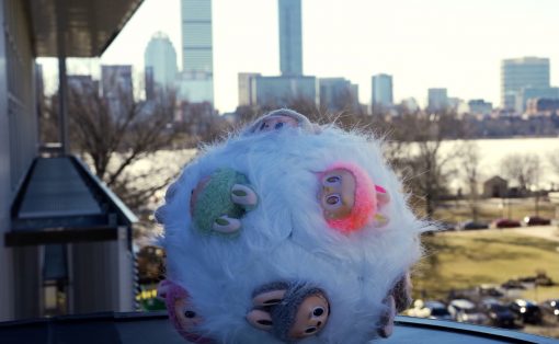 Fluffy white plush with colorful monster patches on a rooftop ledge, city skyline in the background.