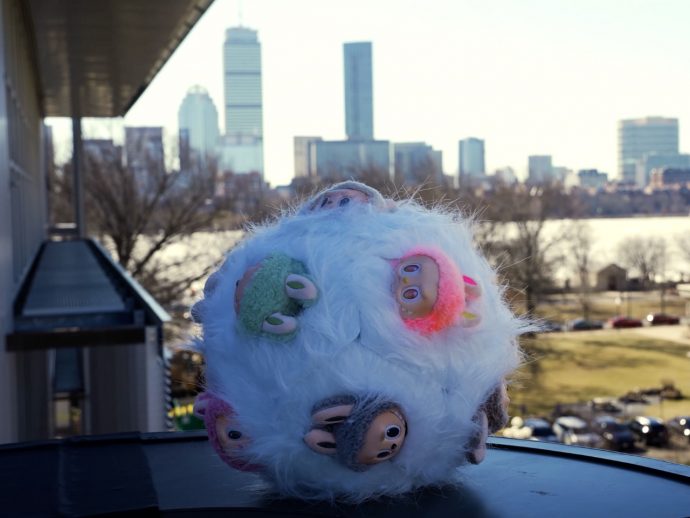 Fluffy white plush with colorful monster patches on a rooftop ledge, city skyline in the background.