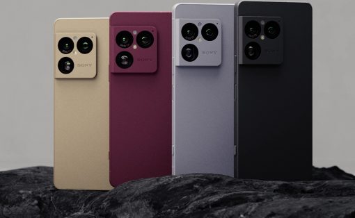 Four smartphones in beige, burgundy, gray, and black with large quad-camera modules on the back, standing on rocks.”