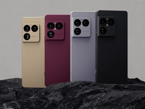 Four smartphones in beige, burgundy, gray, and black with large quad-camera modules on the back, standing on rocks.”