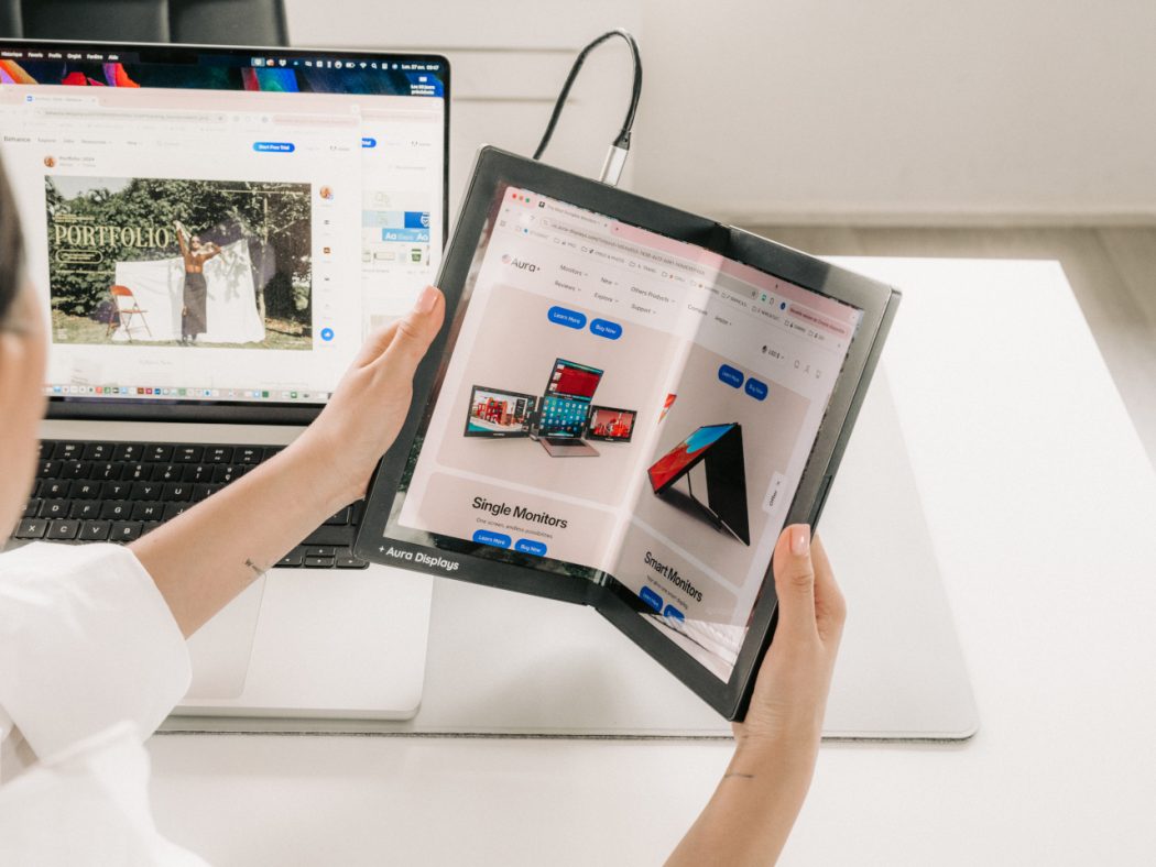 Person holds a foldable tablet opened to a product catalog beside a laptop on a white desk