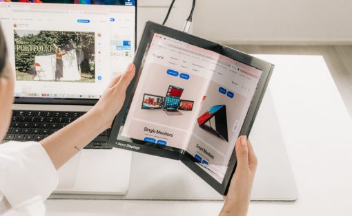 Person holds a foldable tablet opened to a product catalog beside a laptop on a white desk