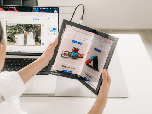 Person holds a foldable tablet opened to a product catalog beside a laptop on a white desk