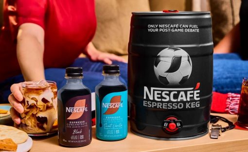 A table with a large NESCAFÉ Espresso Keg, two NESCAFÉ concentrate bottles, and a glass of swirled iced coffee beside snacks, with people in the background at a casual gathering.