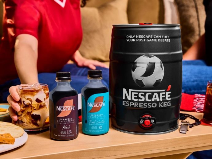 A table with a large NESCAFÉ Espresso Keg, two NESCAFÉ concentrate bottles, and a glass of swirled iced coffee beside snacks, with people in the background at a casual gathering.