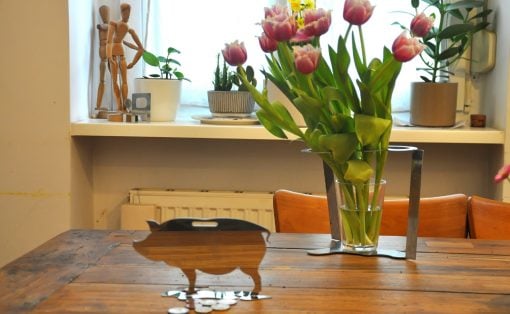 Wooden piggy bank silhouette with coins on a rustic table, beside a vase of pink tulips and potted plants on a bright windowsill.