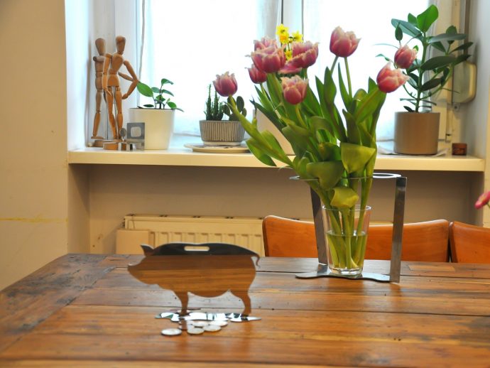 Wooden piggy bank silhouette with coins on a rustic table, beside a vase of pink tulips and potted plants on a bright windowsill.