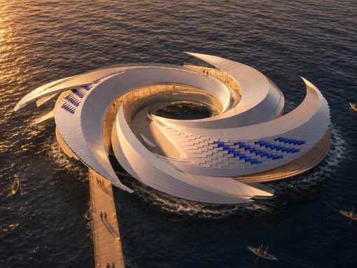 Aerial view of a futuristic white spiral pavilion on the sea, connected by a wooden pier at sunset.