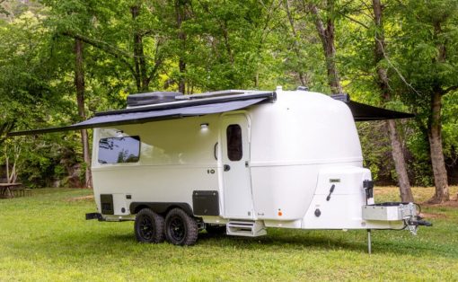 White travel trailer with extended awning parked on grass in a forested campsite.