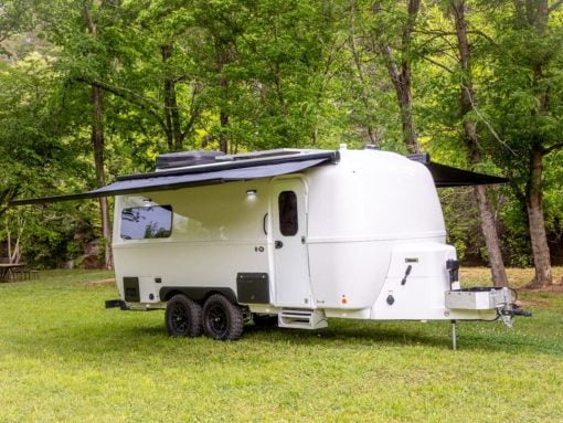 White travel trailer with extended awning parked on grass in a forested campsite.