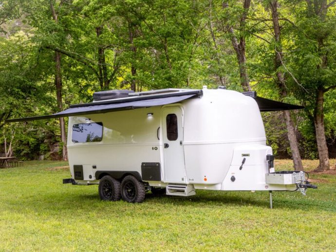 White travel trailer with extended awning parked on grass in a forested campsite.