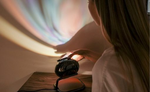 Person adjusting a black decorative projection lamp on a wooden table, casting pastel colors on the wall behind.