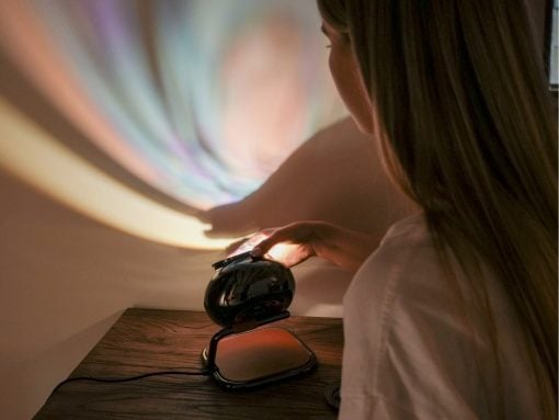 Person adjusting a black decorative projection lamp on a wooden table, casting pastel colors on the wall behind.