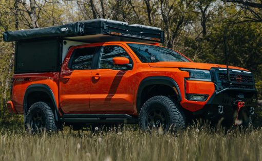 Orange off-road pickup truck with a rooftop camper and gear in a grassy field with trees in the background.