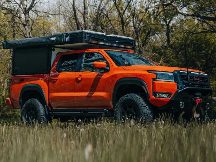 Orange off-road pickup truck with a rooftop camper and gear in a grassy field with trees in the background.