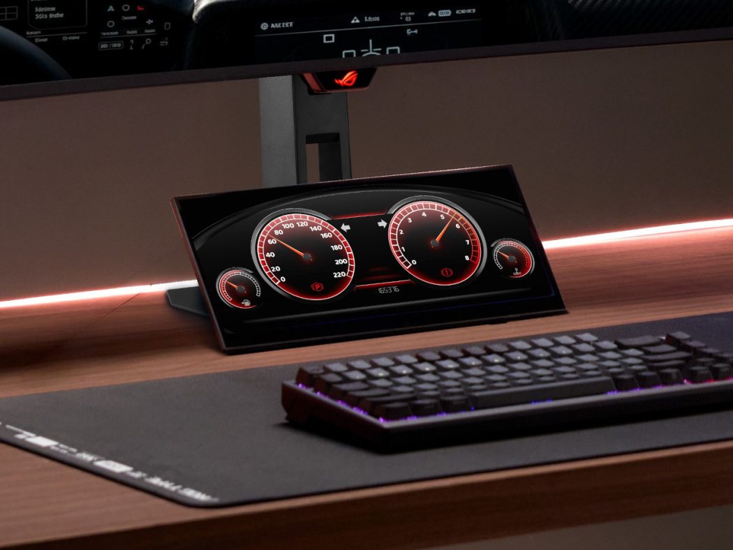 Angled monitor displaying a red-dial car dashboard on a desk, with a black keyboard and large mouse pad nearby.