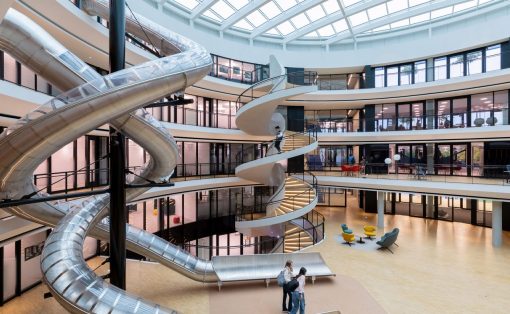 Inside a bright modern atrium with a large spiraling metal slide wrapping around a central column and balconies on multiple levels above.