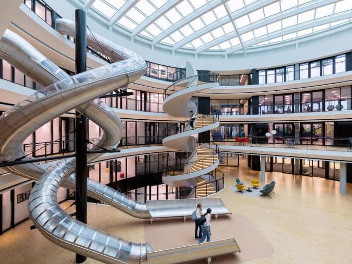 Inside a bright modern atrium with a large spiraling metal slide wrapping around a central column and balconies on multiple levels above.