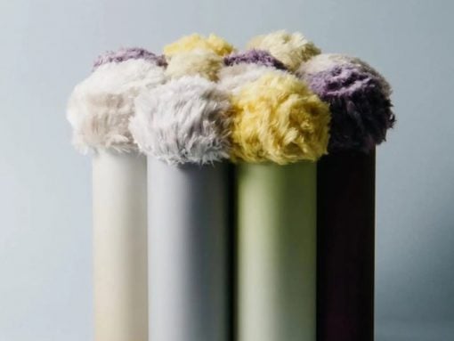Row of pastel-colored cylinders with fluffy yarn pom-poms arranged on top, forming a soft still life.