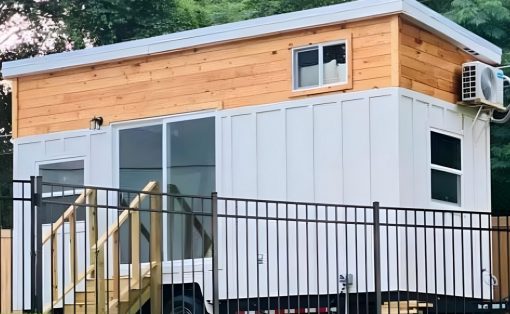 Tiny house on wheels with white siding and a wooden upper level, behind a black metal fence.
