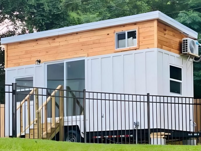 Tiny house on wheels with white siding and a wooden upper level, behind a black metal fence.