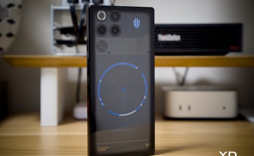 Smartphone standing upright on a wooden desk displaying a circular blue progress graphic on the screen, with blurred computer hardware in the background.