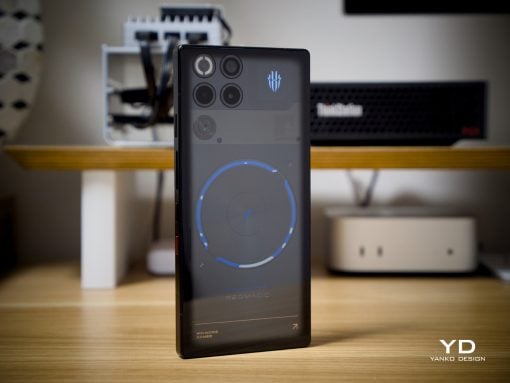 Smartphone standing upright on a wooden desk displaying a circular blue progress graphic on the screen, with blurred computer hardware in the background.
