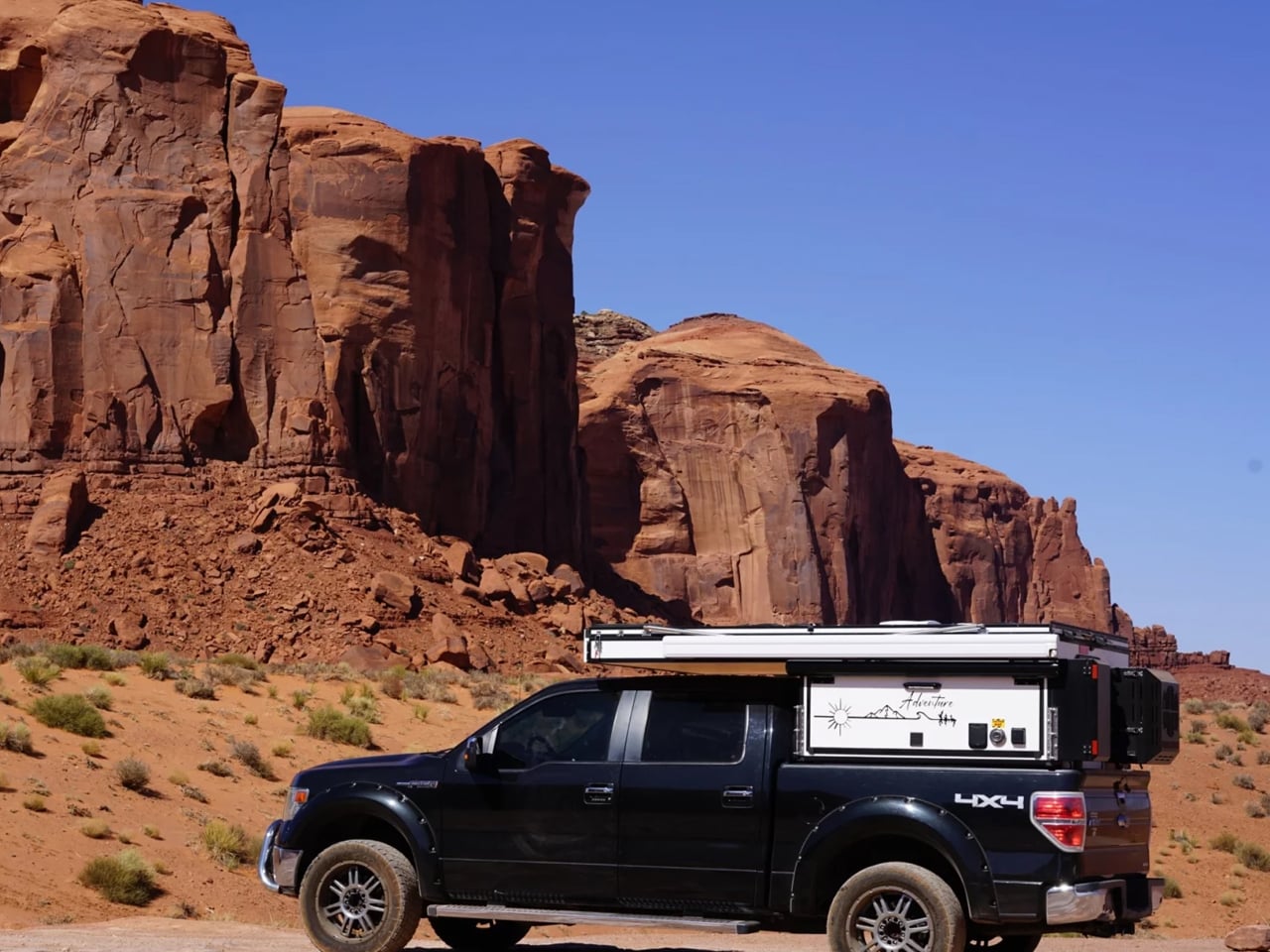 Black pickup truck with a rooftop camper parked in a desert landscape of red rock formations under a blue sky.