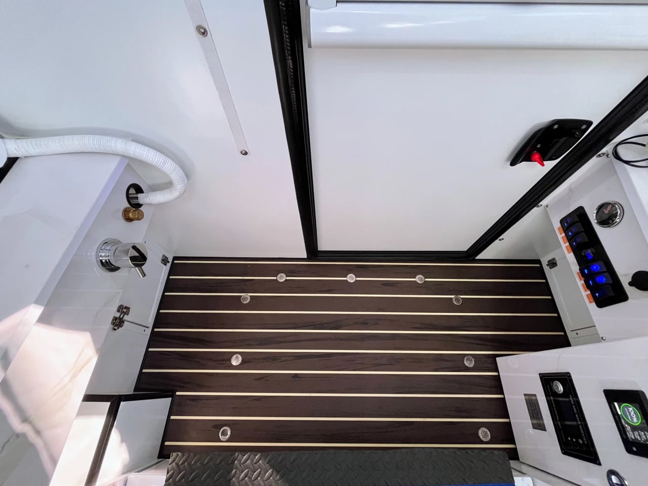 Interior of a compact vehicle bathroom with dark wood plank flooring, a white sliding door, and a blue-lit control panel on the right.