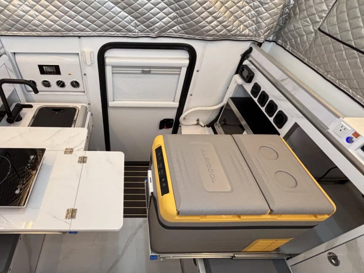 Camper van kitchenette with a fold-out marble counter, sink, stove, and a yellow Alpicool portable cooler on the floor beside the counter.