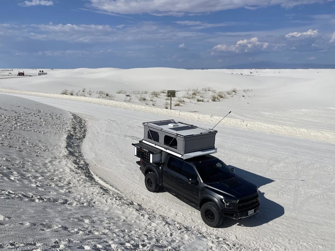 Black Ford pickup with a rooftop tent parked on white sand dunes under a blue sky, a desert campsite setup on the vehicle's bed.