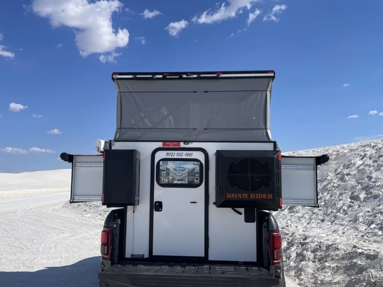 White pickup with a raised rooftop tent against a bright blue sky and white snow dunes behind.