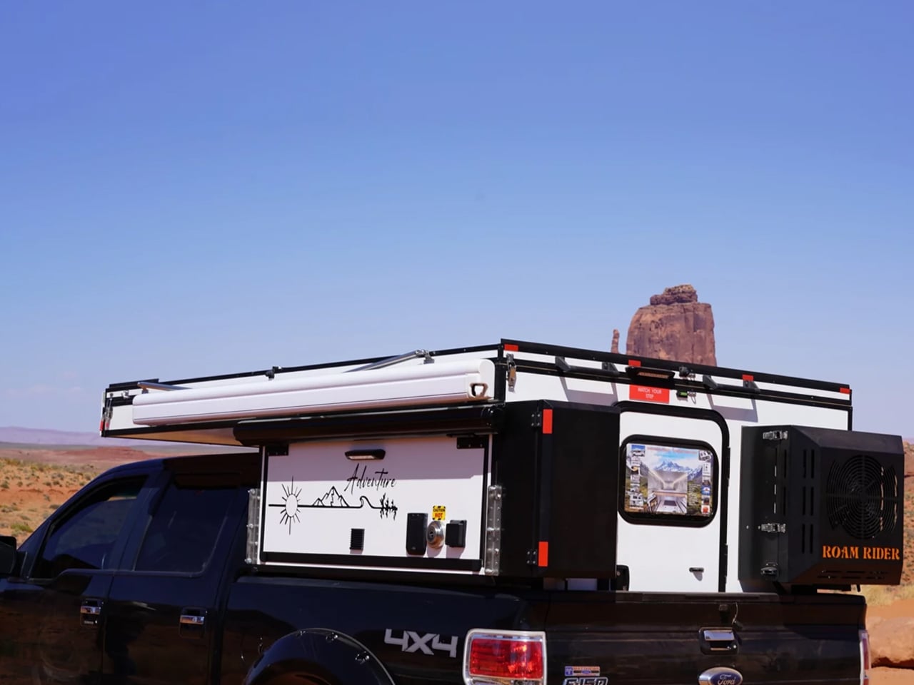 Black pickup truck carrying a white camper shell with rooftop gear in a desert landscape, rock formation in the distance