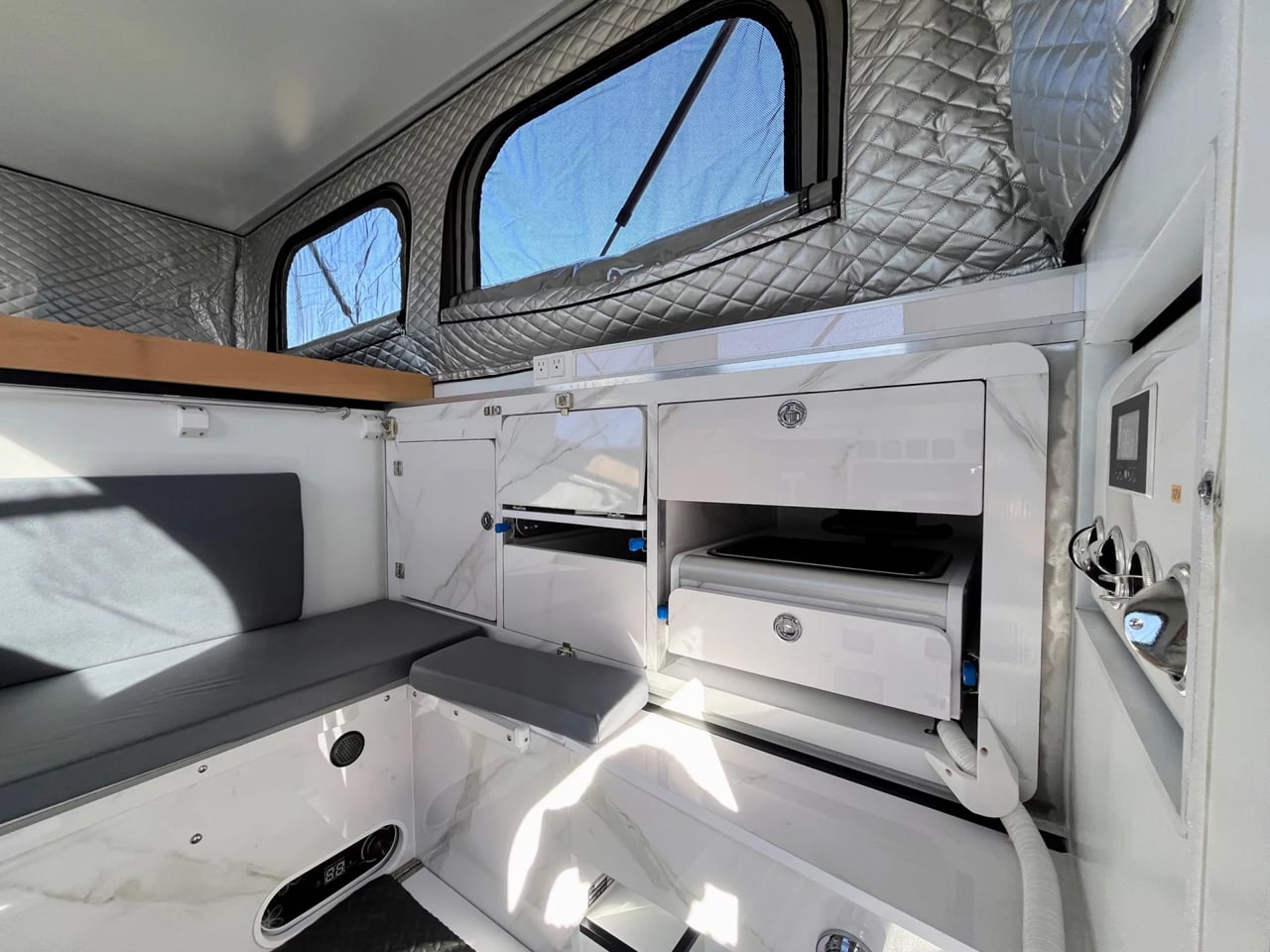 Camper van interior with white marble-look cabinets, a small fridge and drawer, grey cushioned seating, and quilted insulation above windows; compact kitchenette area on right side.