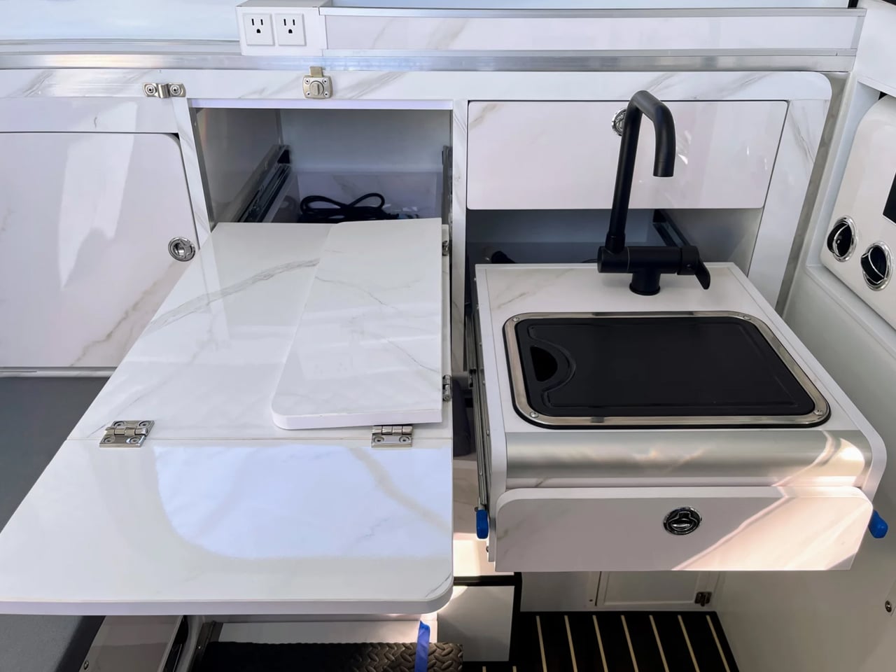 White marble galley with a fold-out counter extension, open storage underneath, and a built-in grill with a black gooseneck faucet on the right side.