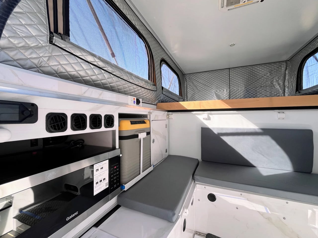 Interior of a compact camper van with a kitchenette, grey cushioned seating, and quilted aluminum walls around windows.