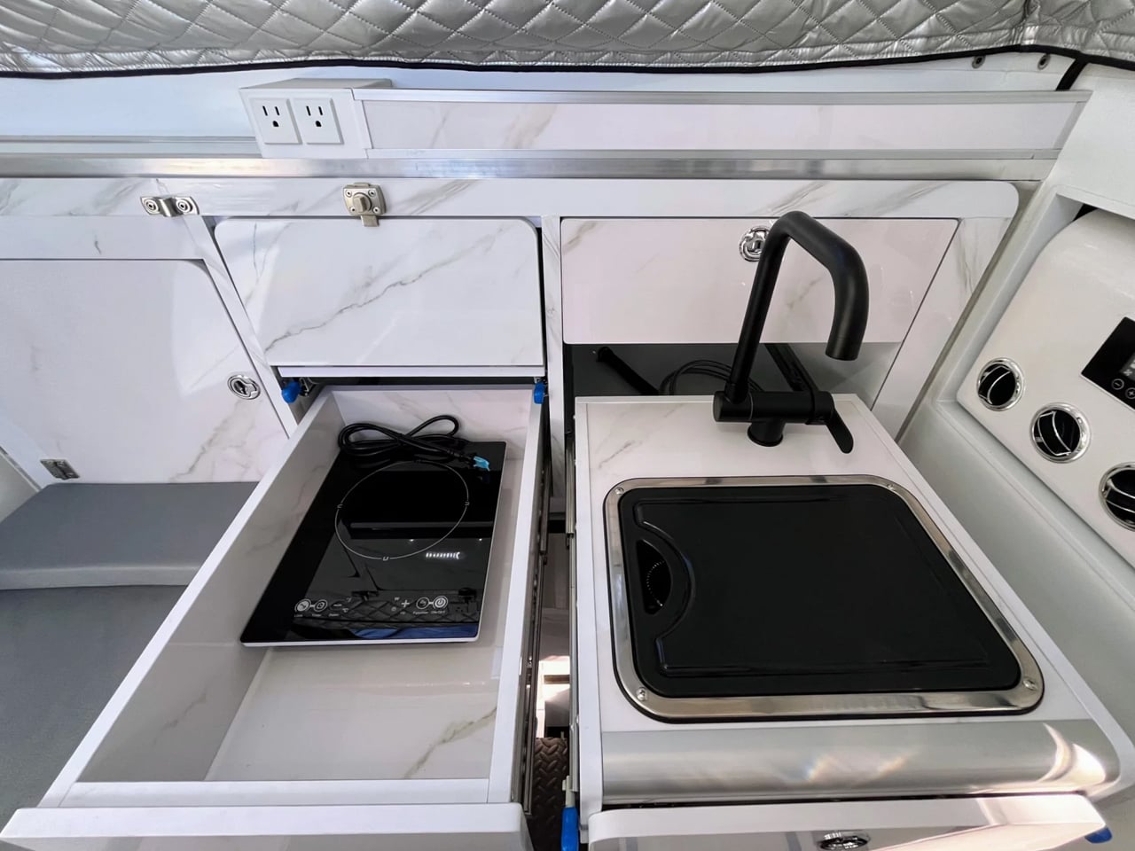 Compact galley aboard a boat or RV with marble counters, a pull-out induction cooktop in a drawer, and a black gooseneck faucet over a sink.