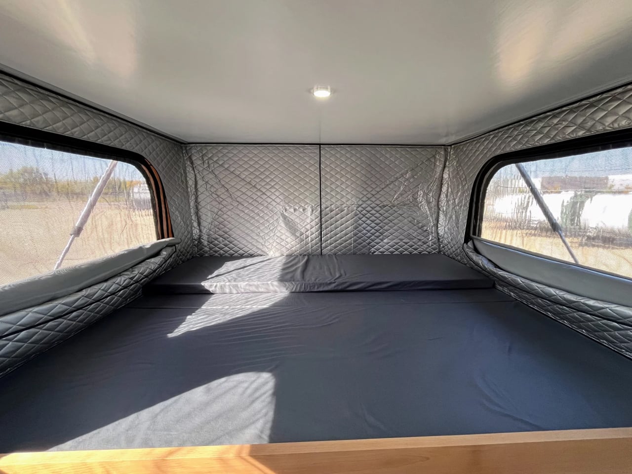 Cozy camper van sleeping area with padded silver quilted walls, a wide gray mattress, and windows on both sides.