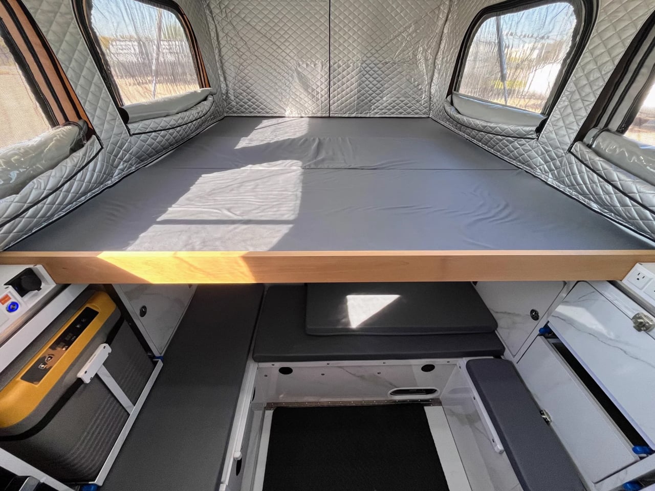Camper van bedroom area with a raised wooden bed frame, grey mattress, and quilted silver insulation walls with side windows behind pleated blinds.