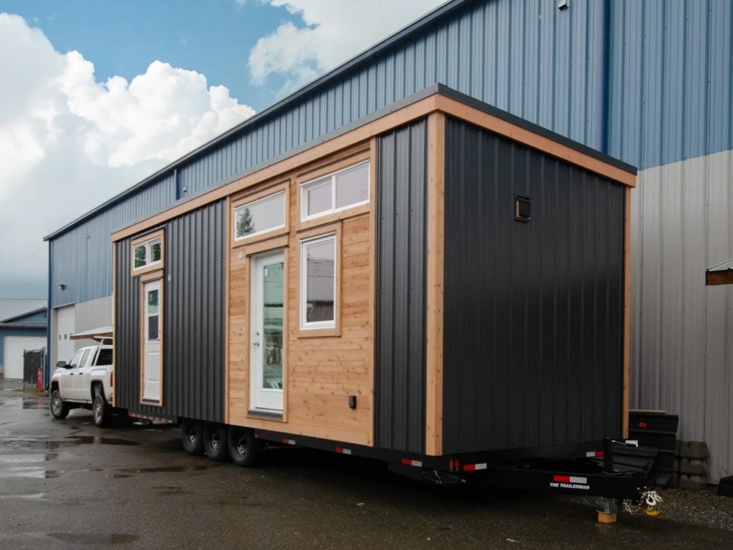 Modular tiny house on a dual-axle trailer, parked beside a blue industrial building with metal siding.