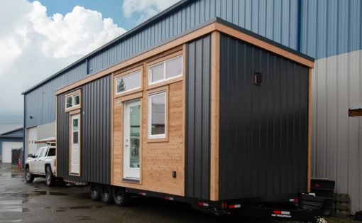 Modular tiny house on a dual-axle trailer, parked beside a blue industrial building with metal siding.