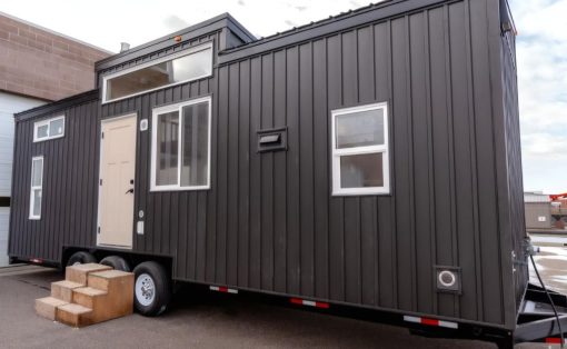 Dark gray modular tiny house on a trailer with a beige front door, large window, and small wooden steps leading up to the entrance by the wheels.