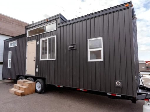 Dark gray modular tiny house on a trailer with a beige front door, large window, and small wooden steps leading up to the entrance by the wheels.
