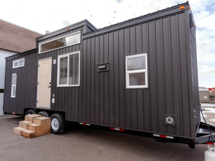 Dark gray modular tiny house on a trailer with a beige front door, large window, and small wooden steps leading up to the entrance by the wheels.