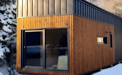 Modern tiny house with wood siding and a metal roof, set in a snowy landscape with a large glass sliding door.