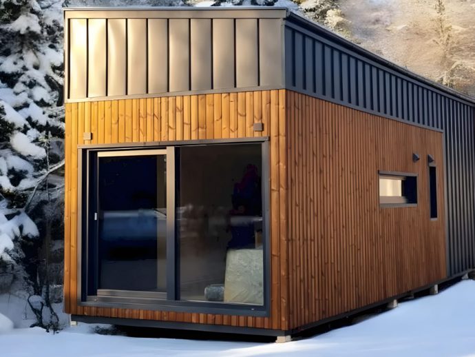Modern tiny house with wood siding and a metal roof, set in a snowy landscape with a large glass sliding door.