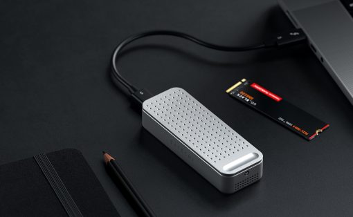 Silver perforated USB-C external drive connected to a laptop on a dark desk, with a pencil and notebook nearby.