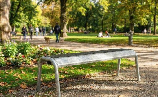 Metal park bench in foreground with a dirt path and autumn leaves; people and trees in a sunny park background.