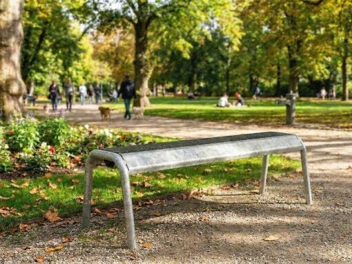 Metal park bench in foreground with a dirt path and autumn leaves; people and trees in a sunny park background.